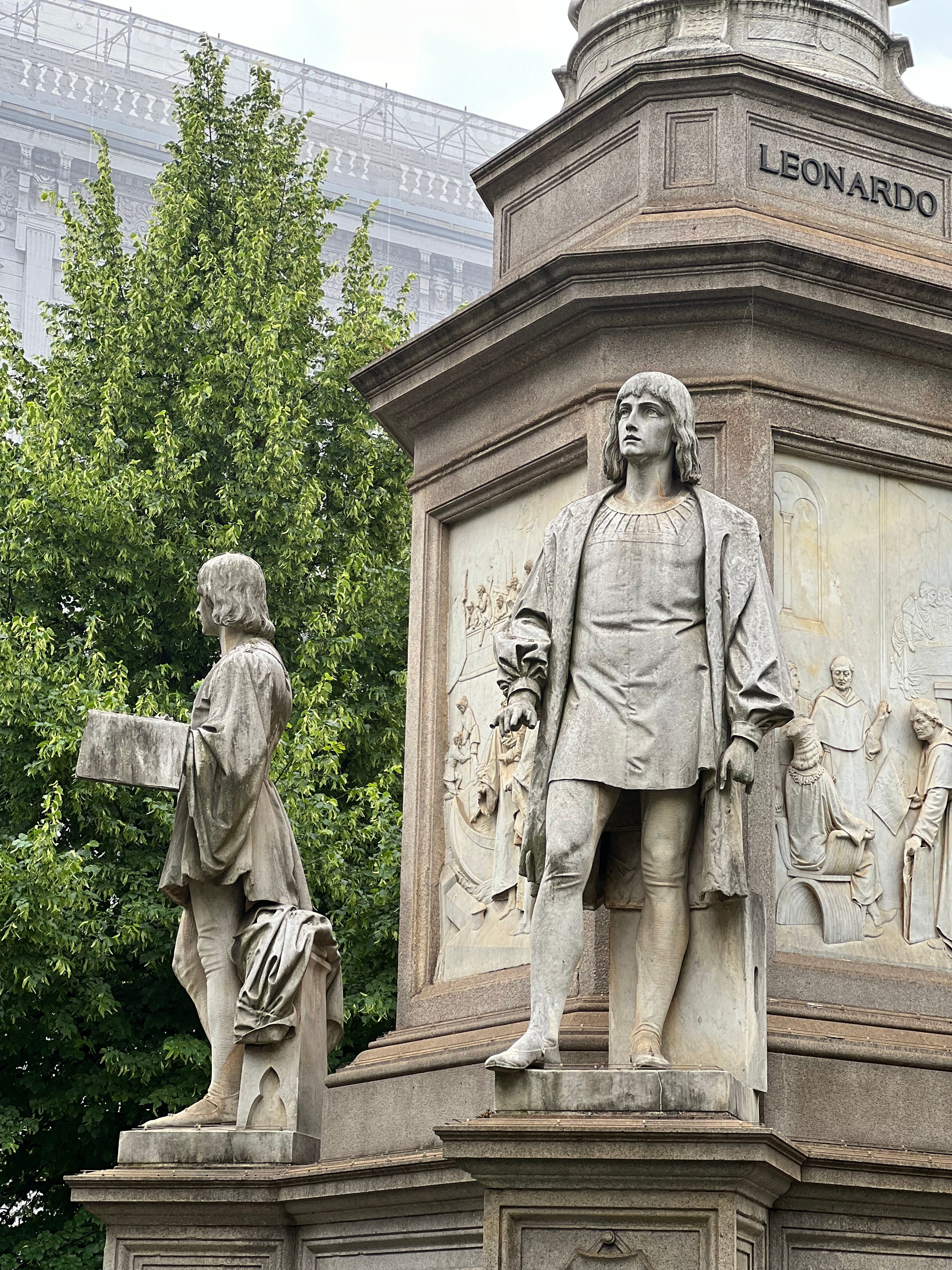 Stone statues of Leonardo da Vinci in a historic monument surrounded by greenery in Milan.
