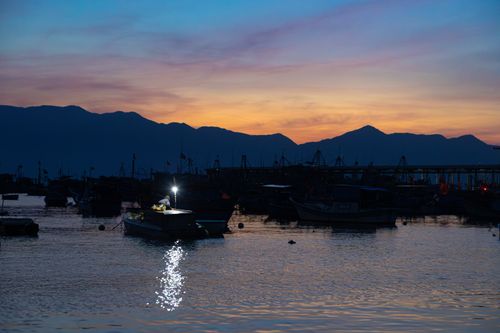 Free A calm harbor scene at twilight with boats silhouetted against a colorful sunset. Stock Photo