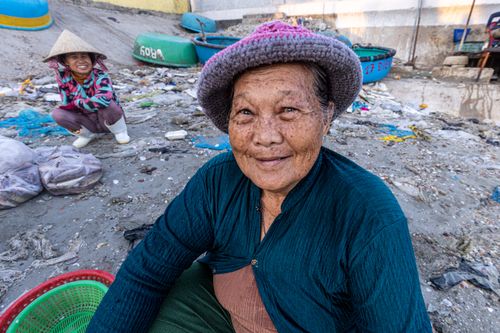 Free Elderly woman smiling on a beach with fishing boats in the background. Vibrant and candid scene. Stock Photo
