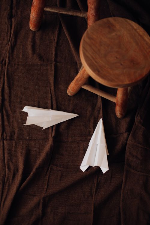 Free Two paper airplanes beside a rustic wooden stool indoors, a brown textile setting. Stock Photo