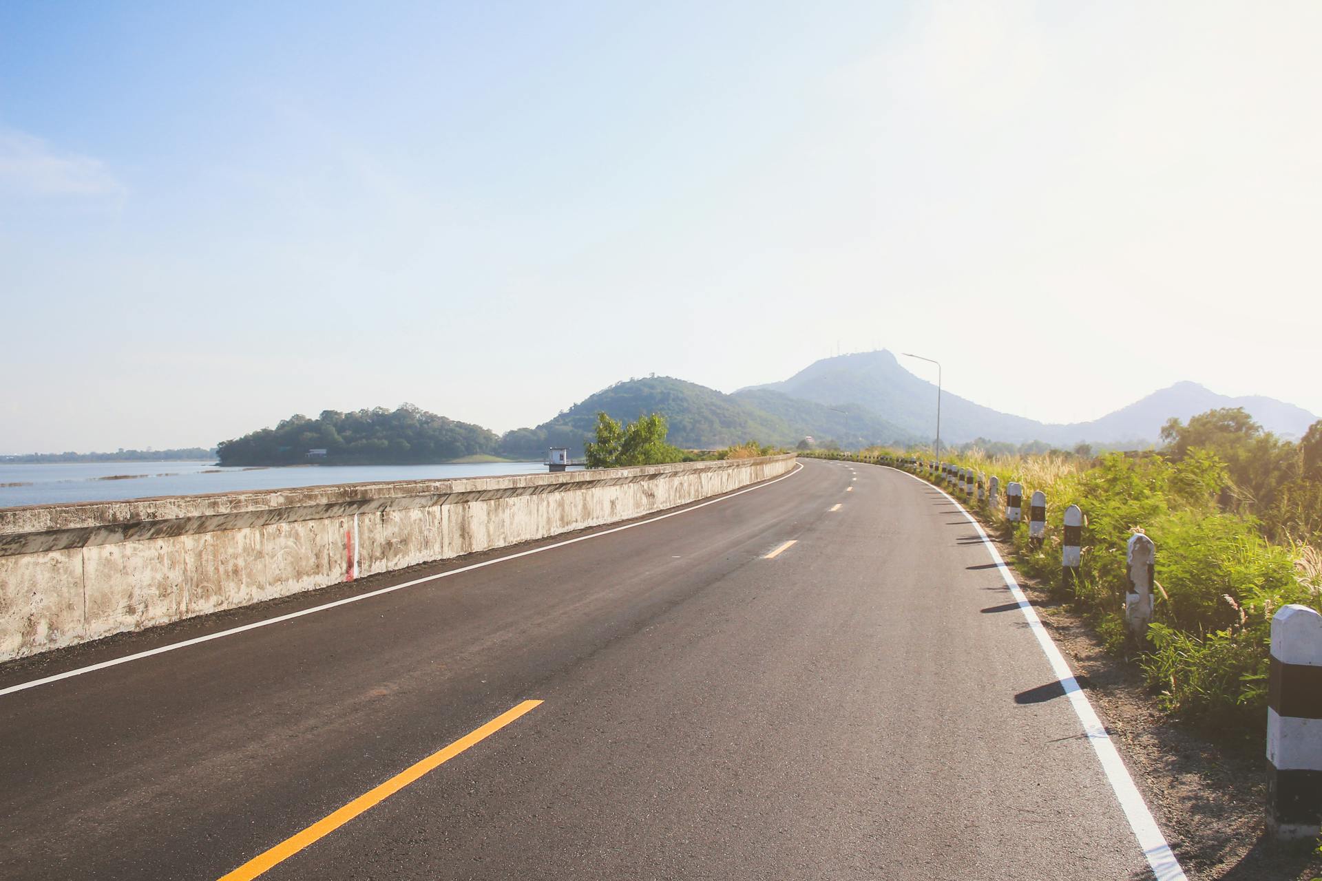 A picturesque empty road curving alongside a tranquil lake, framed by lush mountains under a clear sky.