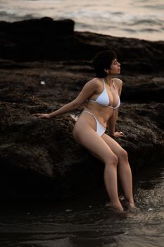 A woman in a white bikini relaxes on seaside rocks during sunset, capturing a serene moment.