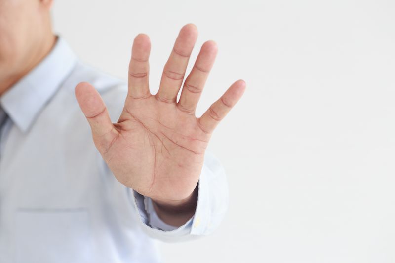 A close-up of an adult male hand extended in a stop gesture against a neutral background.