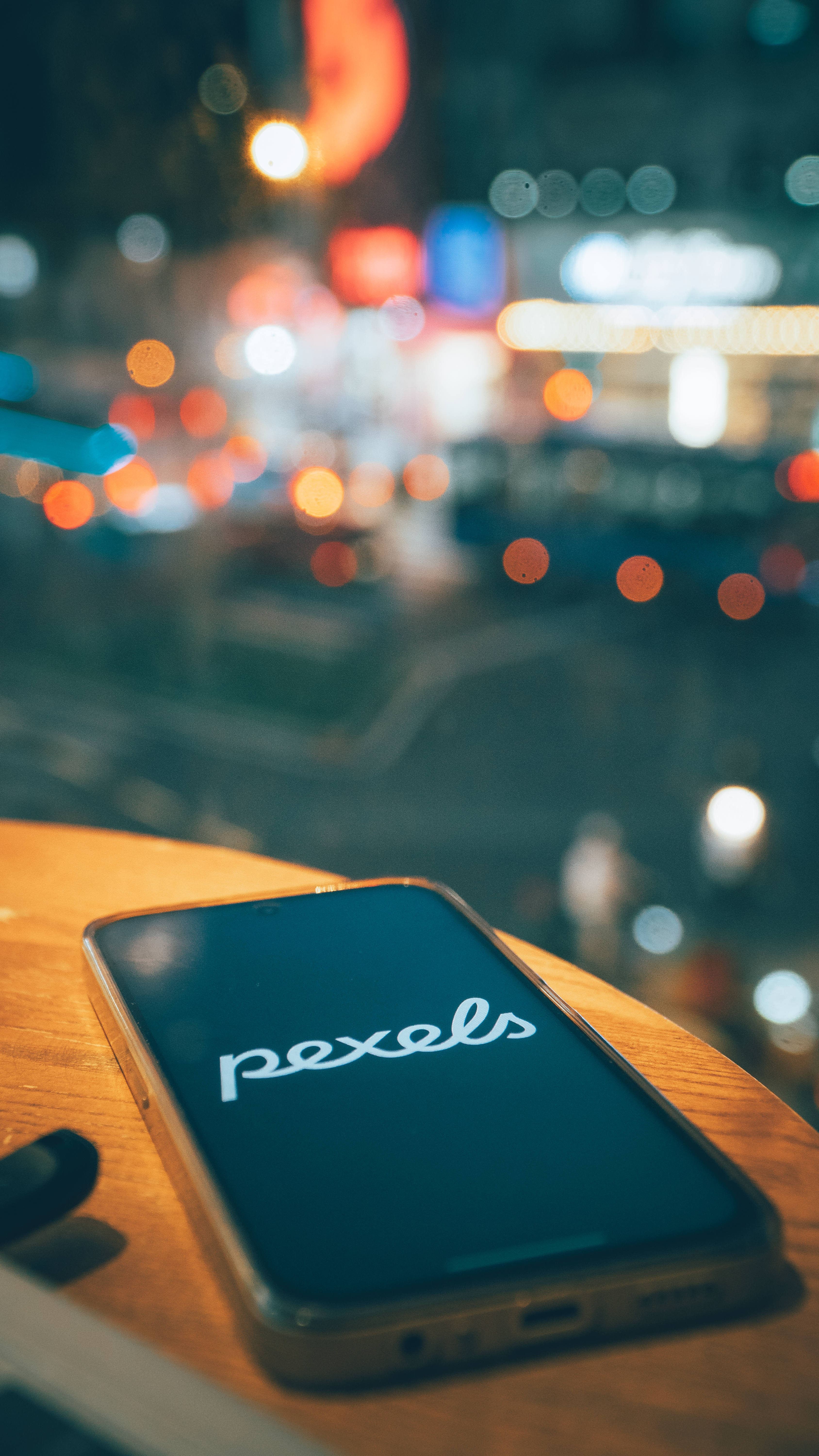 Smartphone with Pexels logo on wooden table, city lights bokeh at night.