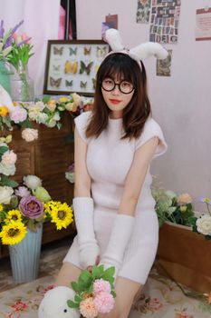 Young woman wearing bunny ears and glasses poses stylishly in a floral-decorated room.
