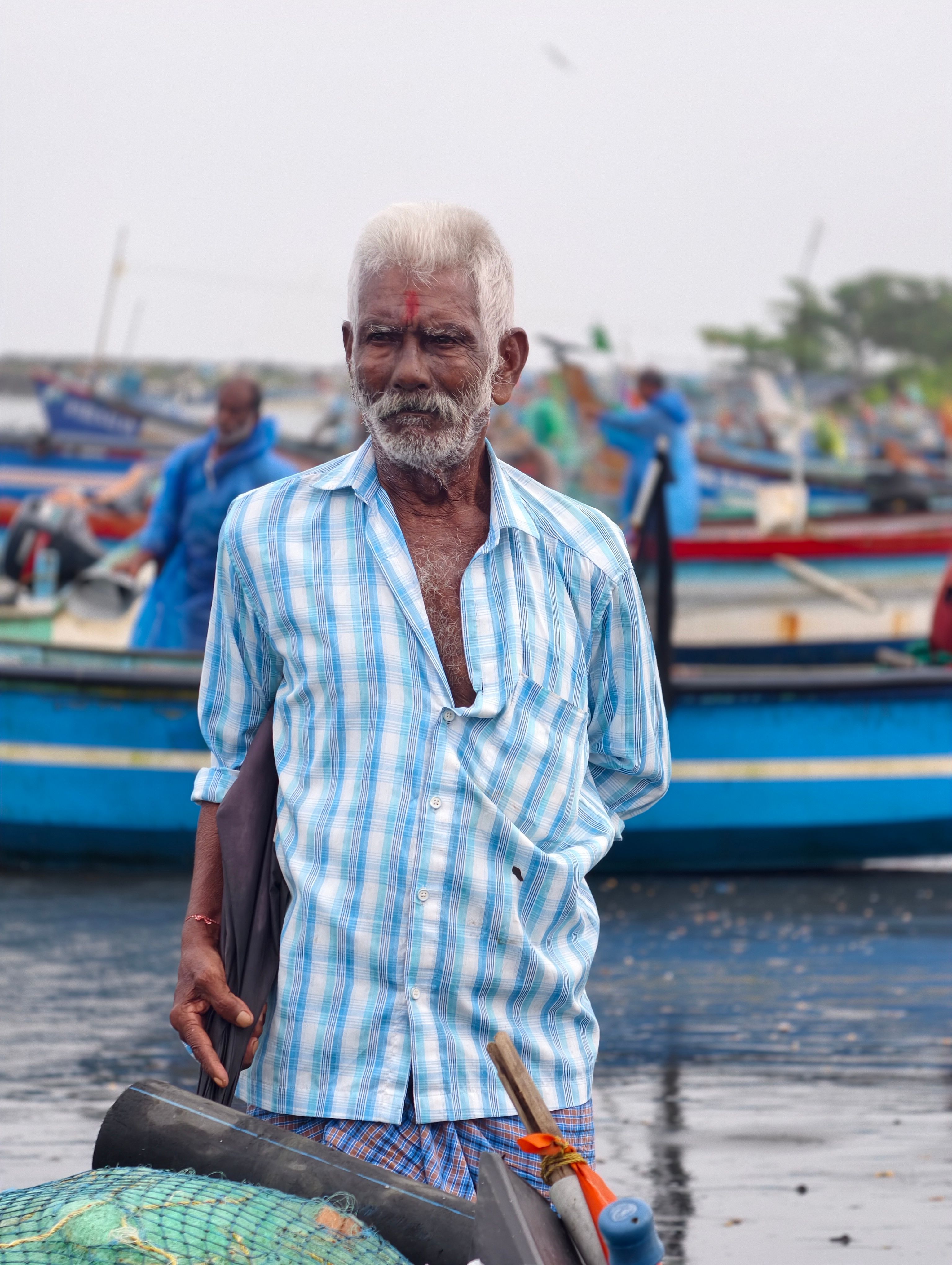 Portrait of an elderly fisherman in a checked shirt standing by colorful boats at a bustling harbor.