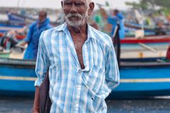 Portrait of an elderly fisherman in a checked shirt standing by colorful boats at a bustling harbor.