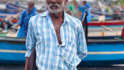 Portrait of an elderly fisherman in a checked shirt standing by colorful boats at a bustling harbor.