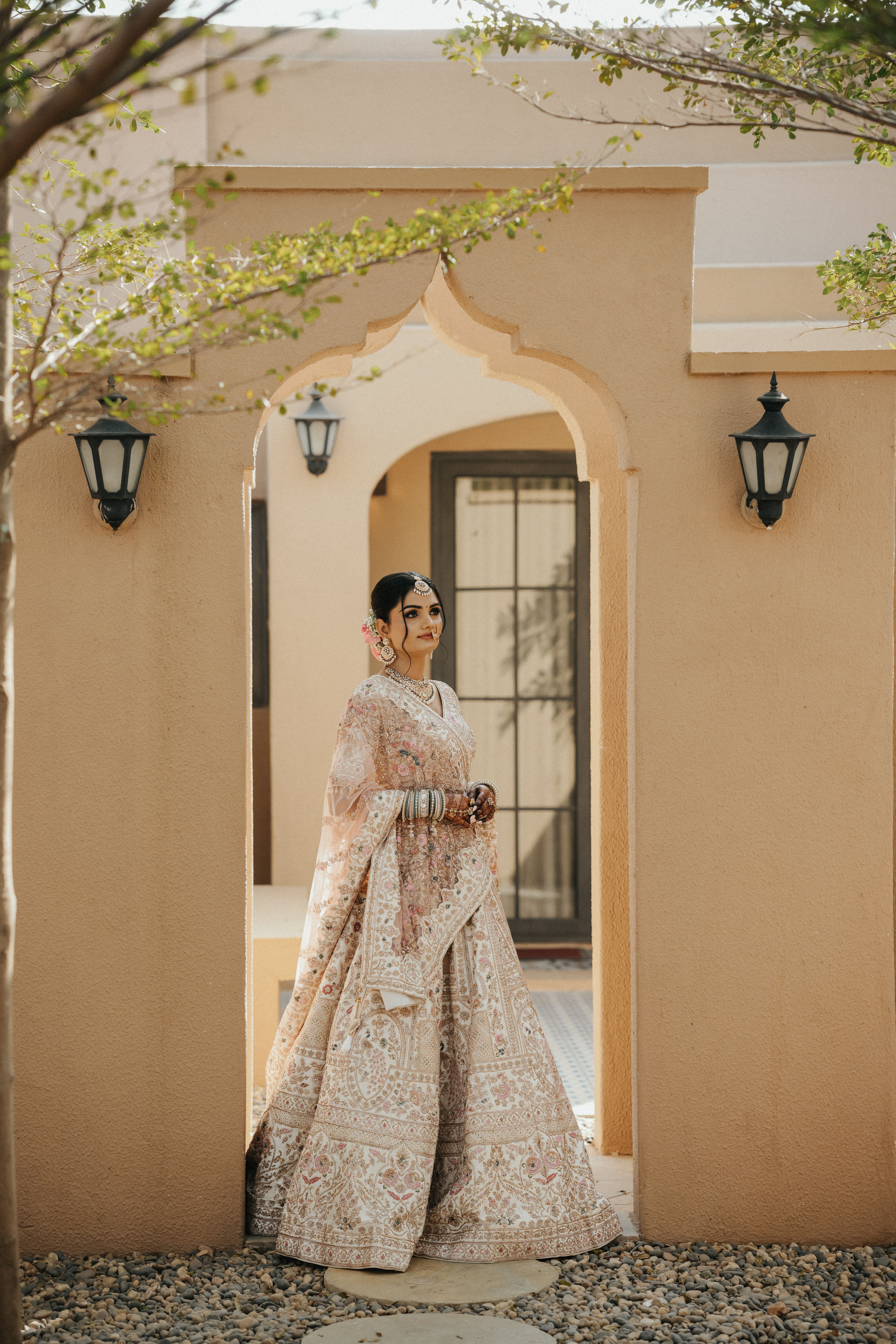 A bride in elaborate traditional attire poses outdoors under an arched doorway, showcasing cultural elegance.