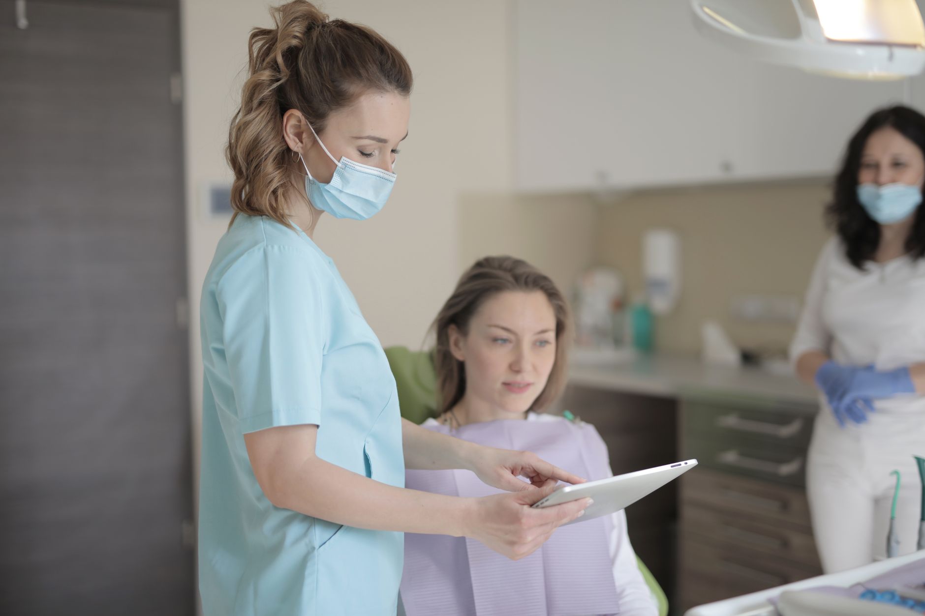 Young female dentist in mask and uniform using tablet while talking with patient before treatment in modern dental clinic in hospital