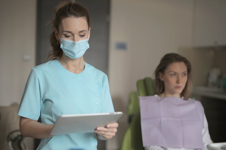 Young Female Dentist In Medical Mask And Uniform Using Tablet Before Treatment Of Patient In Clinic