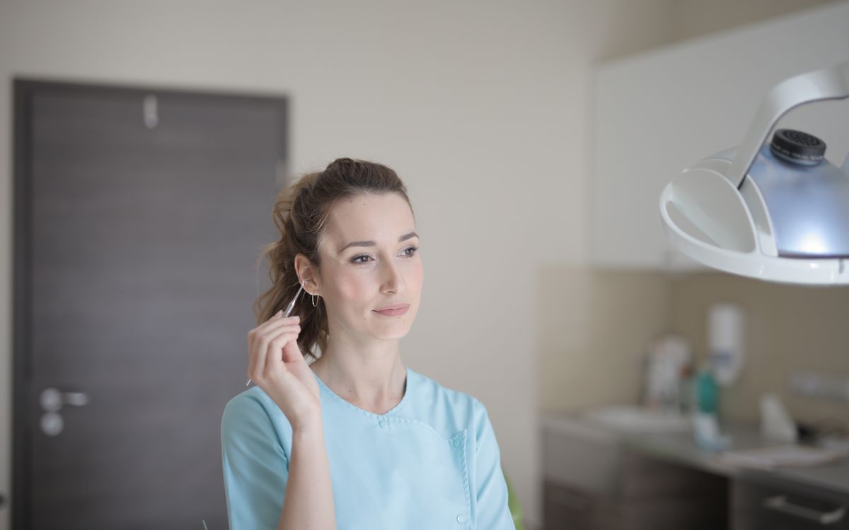 Positive young female doctor wearing blue uniform standing in light modern clinic with metal instruments in hand and looking away thoughtfully