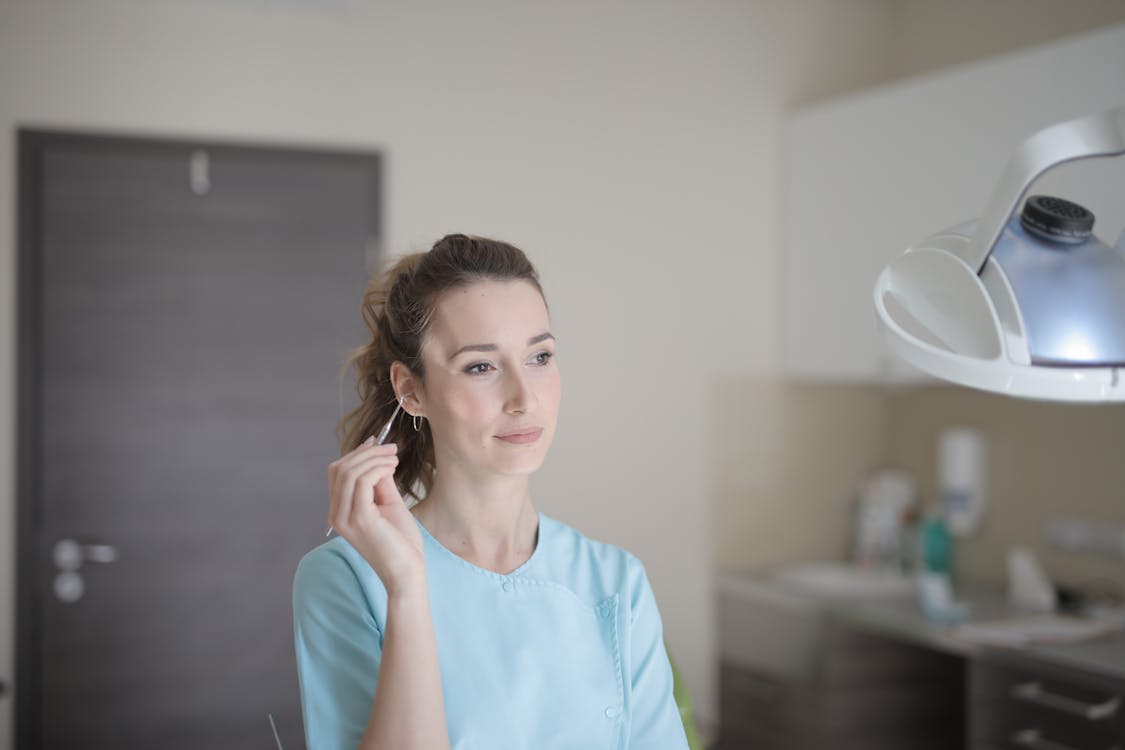 Positive young female doctor wearing blue uniform standing in light modern clinic with metal instruments in hand and looking away thoughtfully