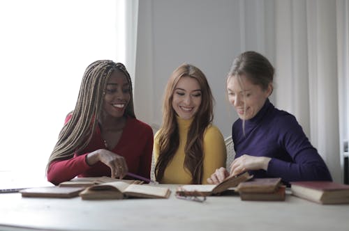 Positive young multiracial women in colorful clothes sitting together at table with books while talking in modern light room