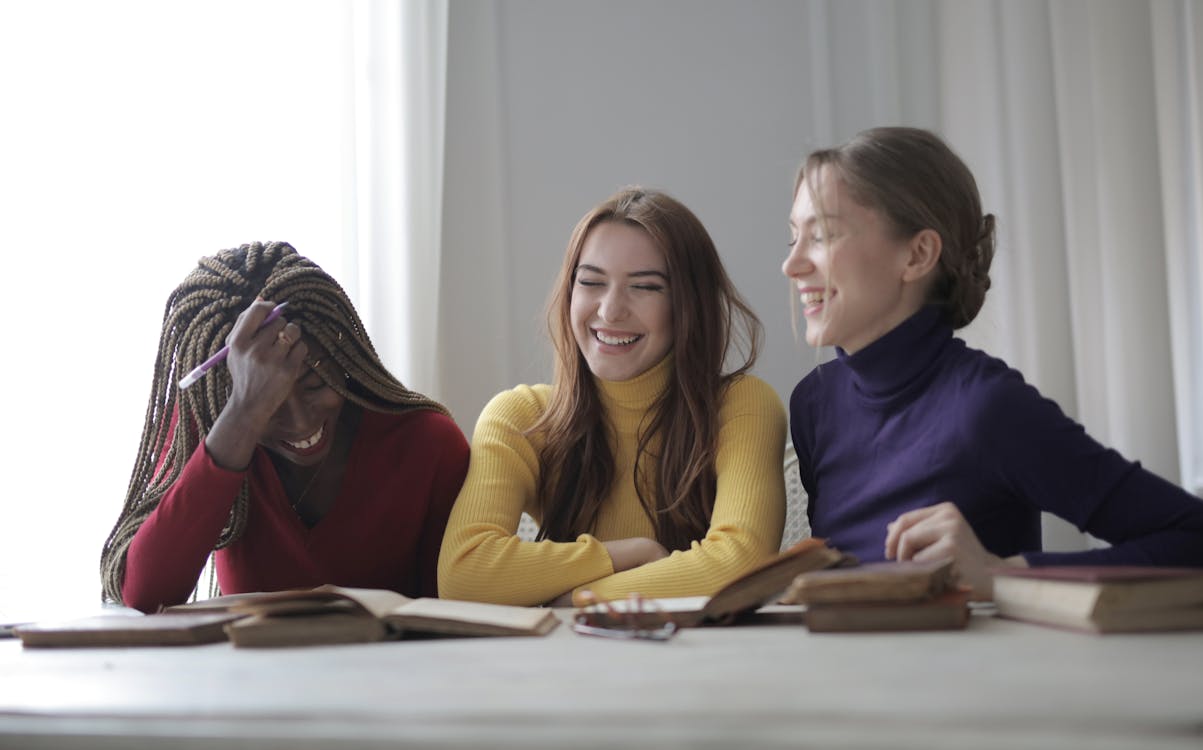 Free Cheerful young multiracial women in colorful casual clothes laughing while sitting together at table with books  in modern light room Stock Photo
