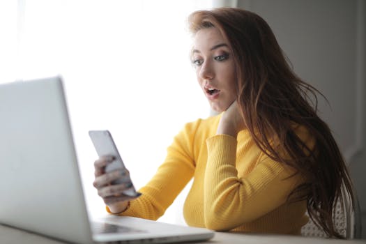 Young woman in yellow sweater looks surprised at phone while working on a laptop indoors.