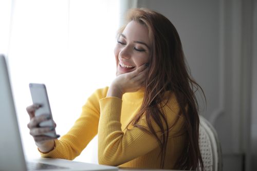 Free Smiling young female freelancer wearing yellow sweater sitting at table near laptop and browsing smartphone while spending time in modern light room Stock Photo