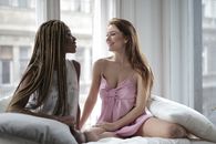 Cheerful young multiethnic girlfriends in pajamas sitting together on comfortable bed near window and looking at each other while spending time in modern apartment
