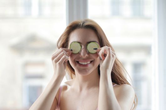Smiling woman with cucumber slices on eyes, enjoying skincare routine indoors.