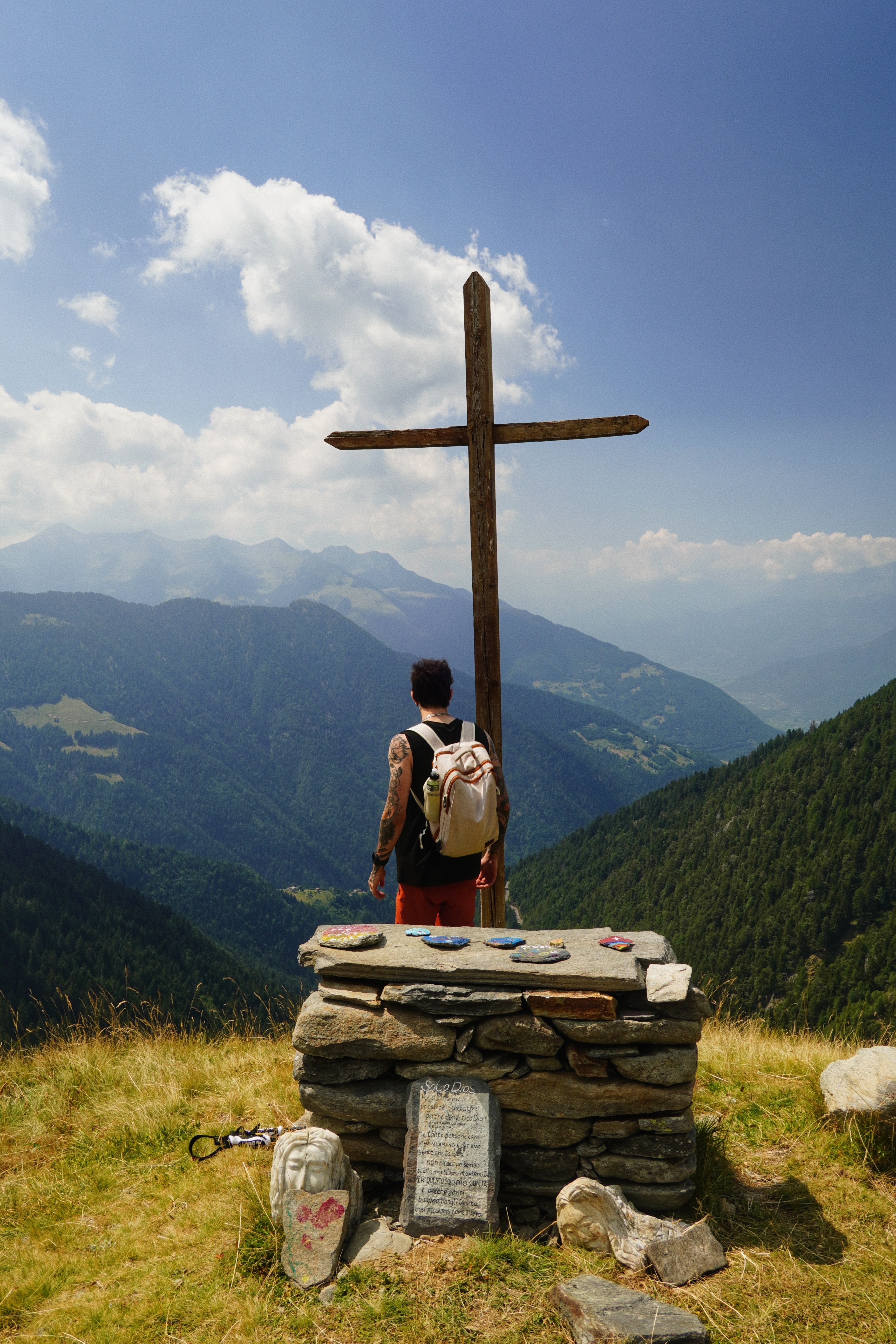 Hiker stands by a cross on a mountain peak, gazing at a scenic valley under blue skies in Lombardia, Italy.