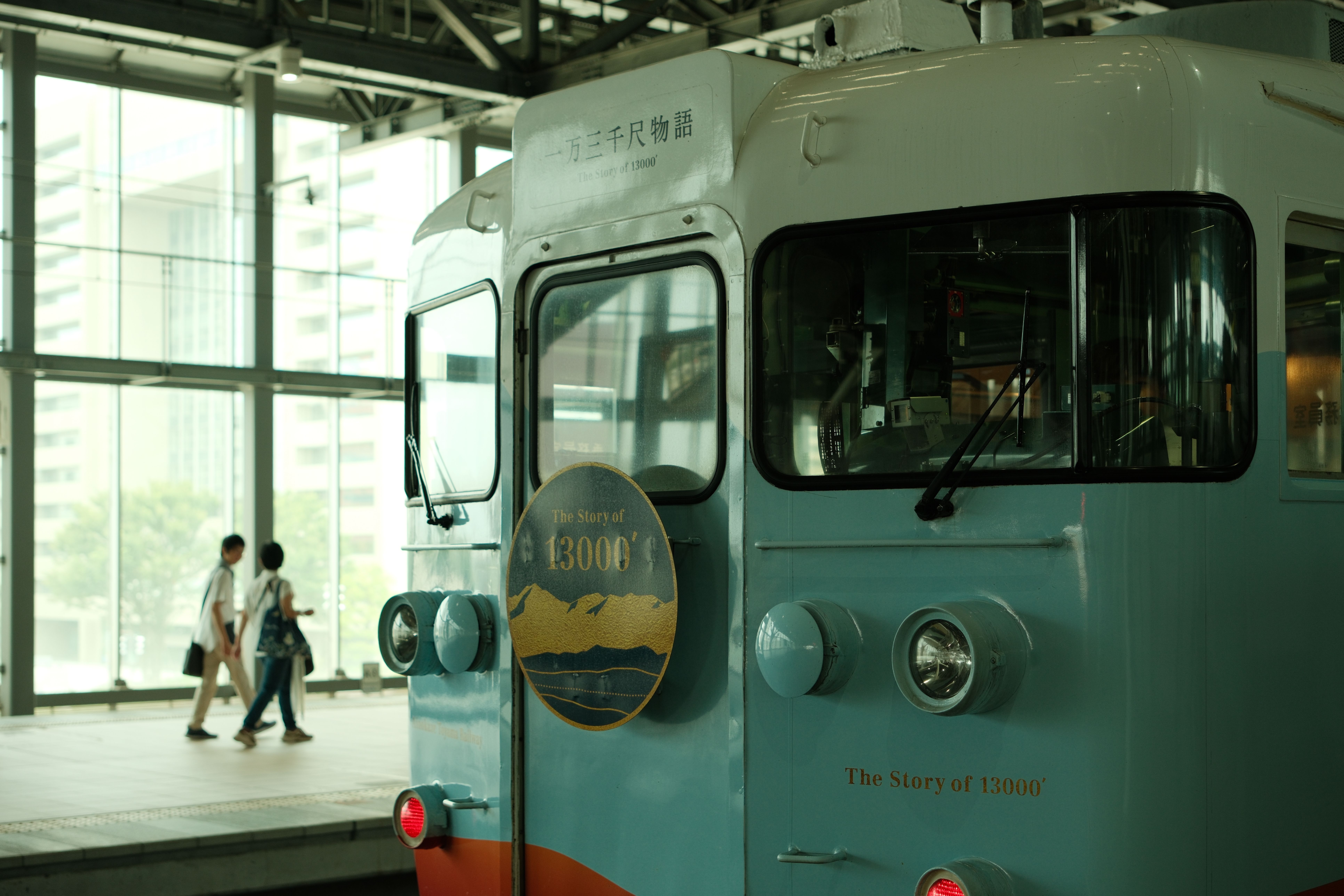 近代的な日本の駅に停車する、クラシックな青い電車。背景には乗客の姿が見える。