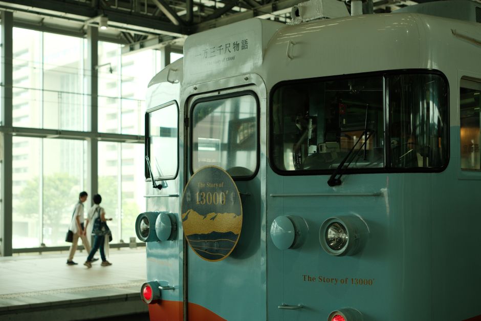 近代的な日本の駅に停車する、クラシックな青い電車。背景には乗客の姿が見える。