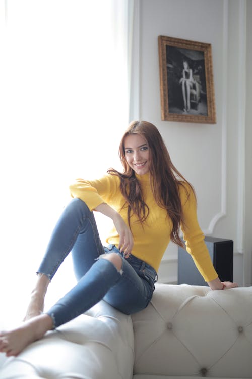 Free Cheerful female in trendy yellow turtleneck and jeans sitting on backrest of chester white sofa against window in classic interior apartment and smiling looking at camera Stock Photo