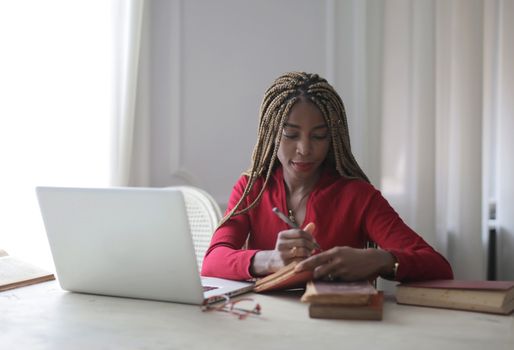 Woman in red shirt writing at desk with laptop, deeply focused.