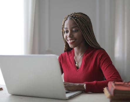 Young black woman happily working on a laptop indoors, showcasing a modern remote work setup.
