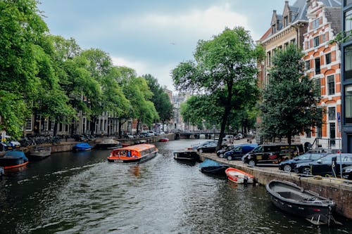 Picturesque scenery of modern vessel floating on calm channel with moored traditional boats amidst beautiful historic buildings and green trees in Amsterdam