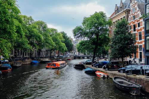 Free Picturesque scenery of modern vessel floating on calm channel with moored traditional boats amidst beautiful historic buildings and green trees in Amsterdam Stock Photo