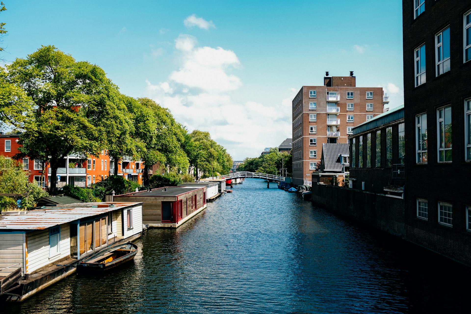Picturesque channel in Amsterdam with traditional houseboats near modern buildings in city outskirts against blue sky