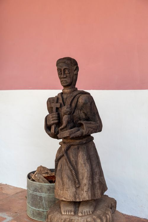 Free Wooden monk sculpture with a cross, placed outdoors against a pink wall. Stock Photo