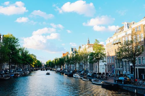 Free Magnificent cityscape of Amsterdam with traditional boats moored on calm river near historic buildings under cloudy blue sky Stock Photo