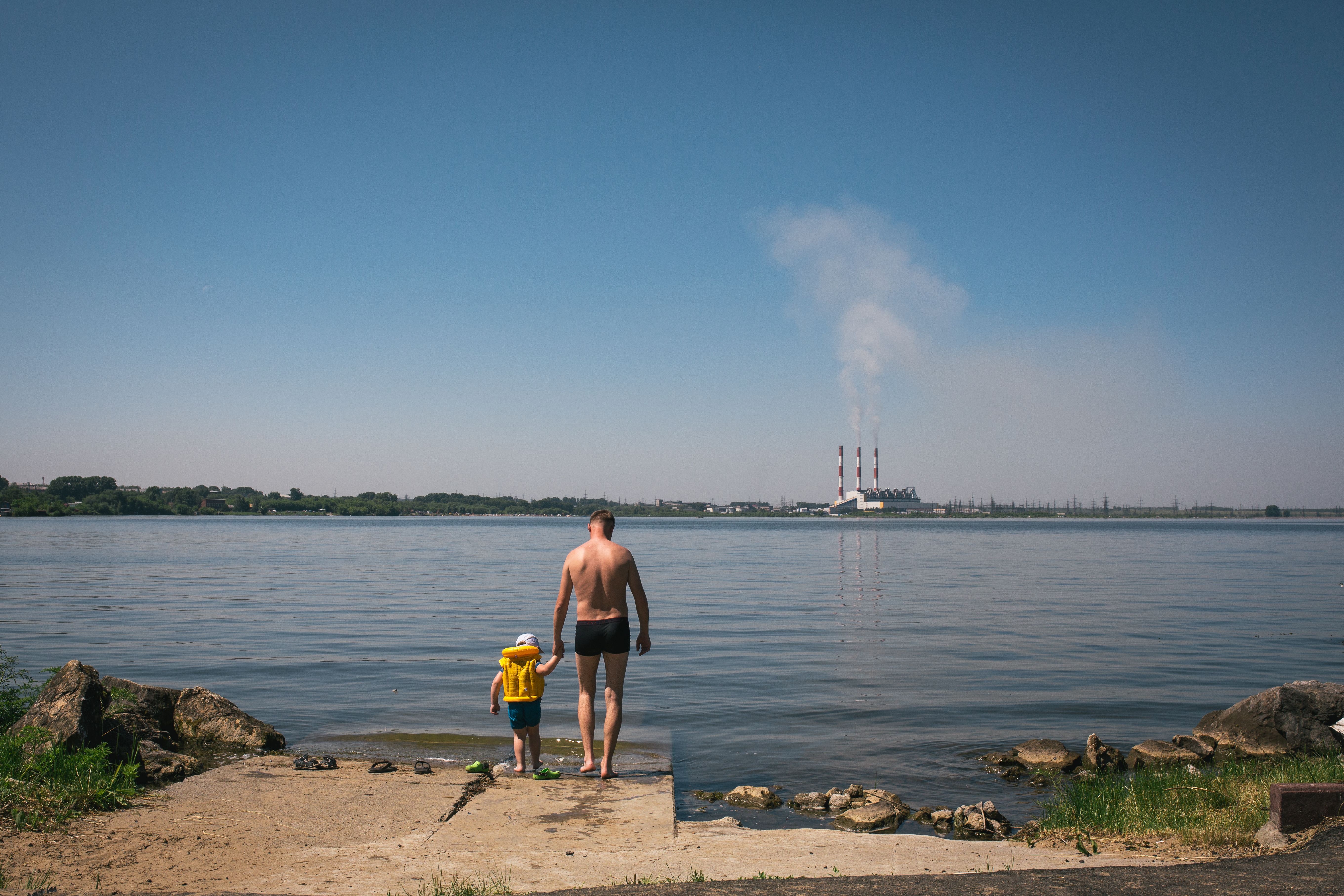 A father and son enjoy a summer day by the lake, with a factory visible in the distance.