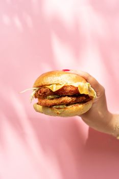 Close-up of a hand holding a crispy chicken burger against a pastel pink background, evoking fresh and modern vibes.