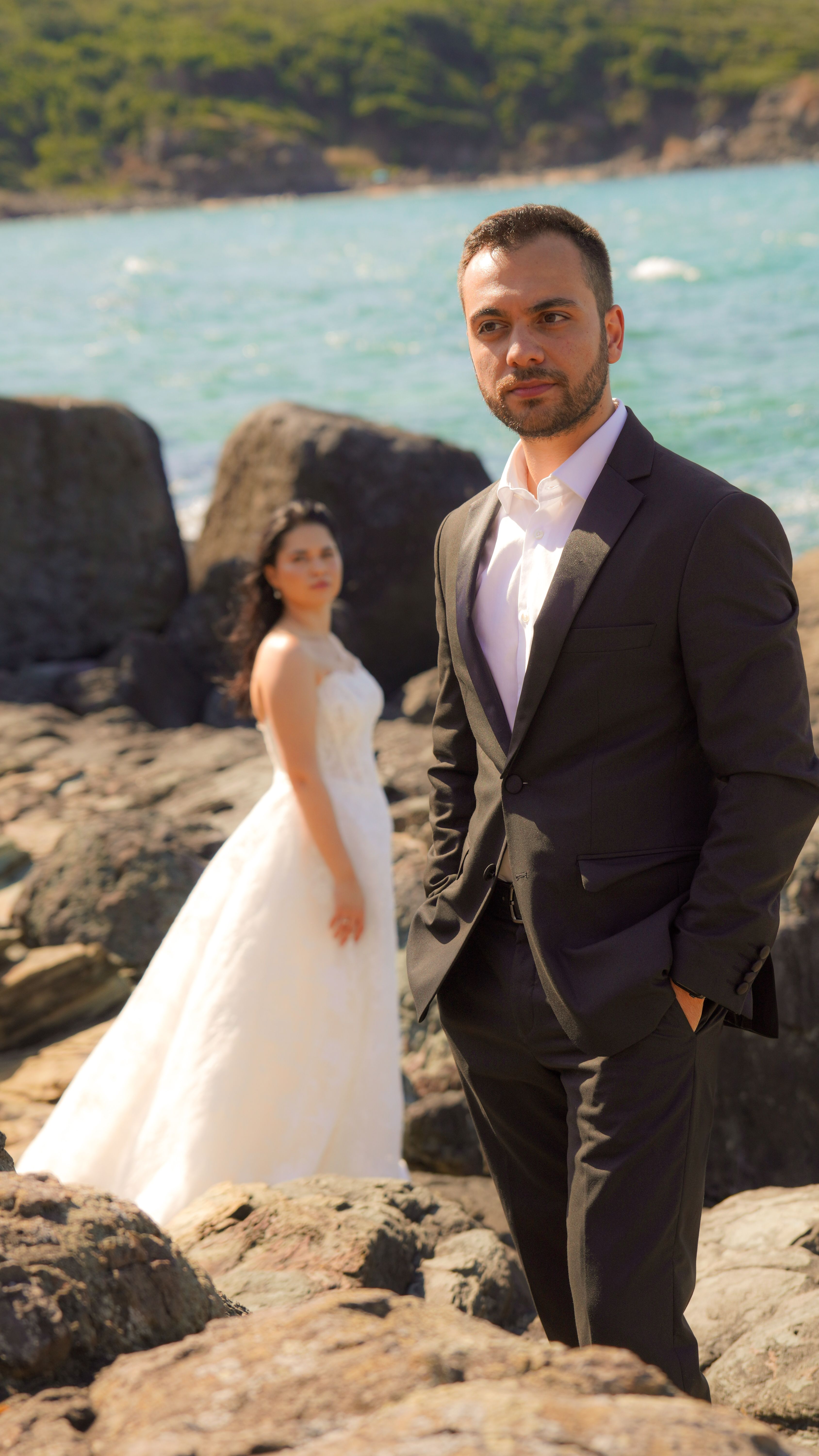 Elegant couple in wedding attire stands on rocky beach by the ocean on a sunny day.