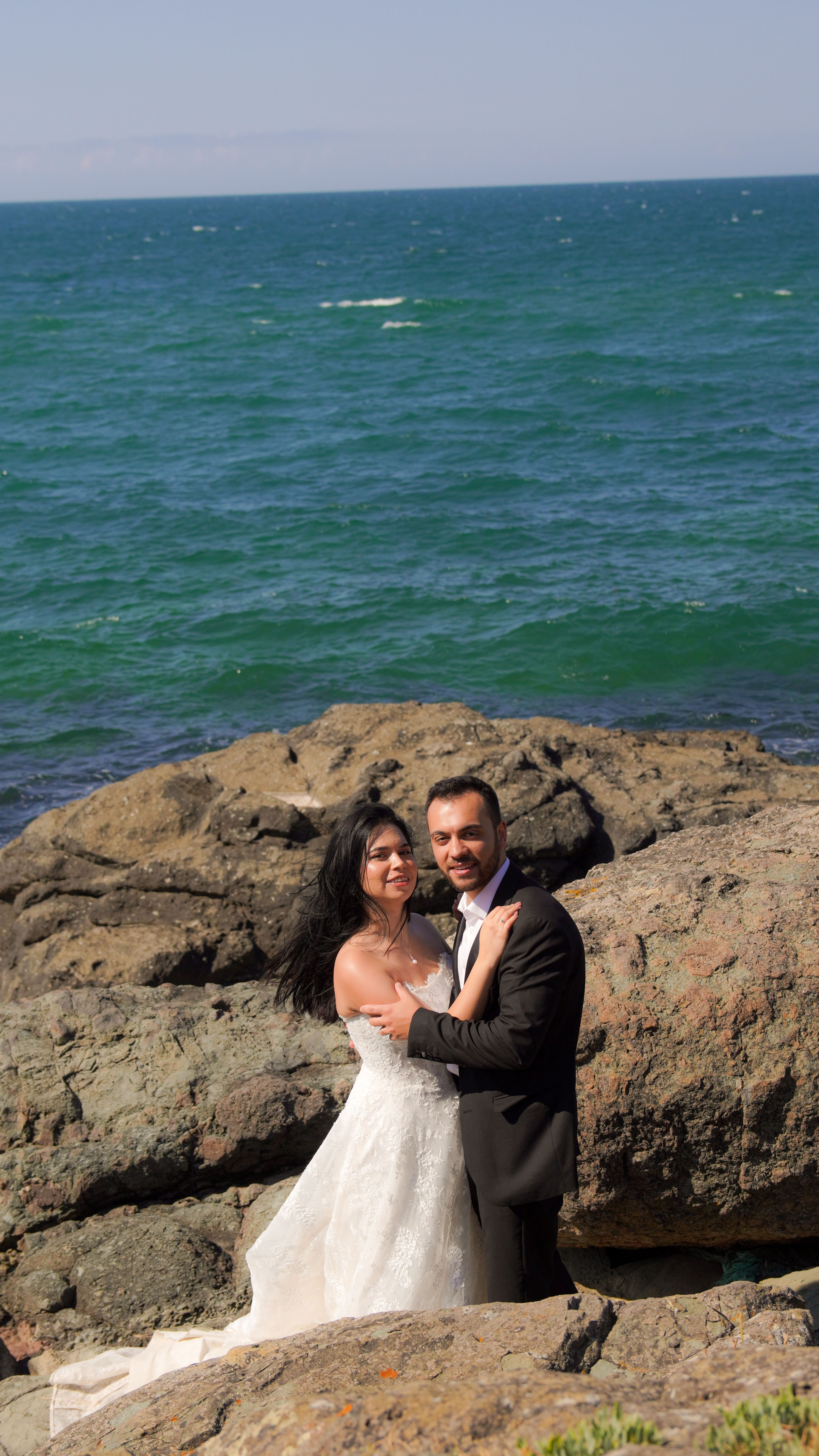 Bride and groom embracing on rocky shore with ocean backdrop, capturing romantic seaside wedding moment.