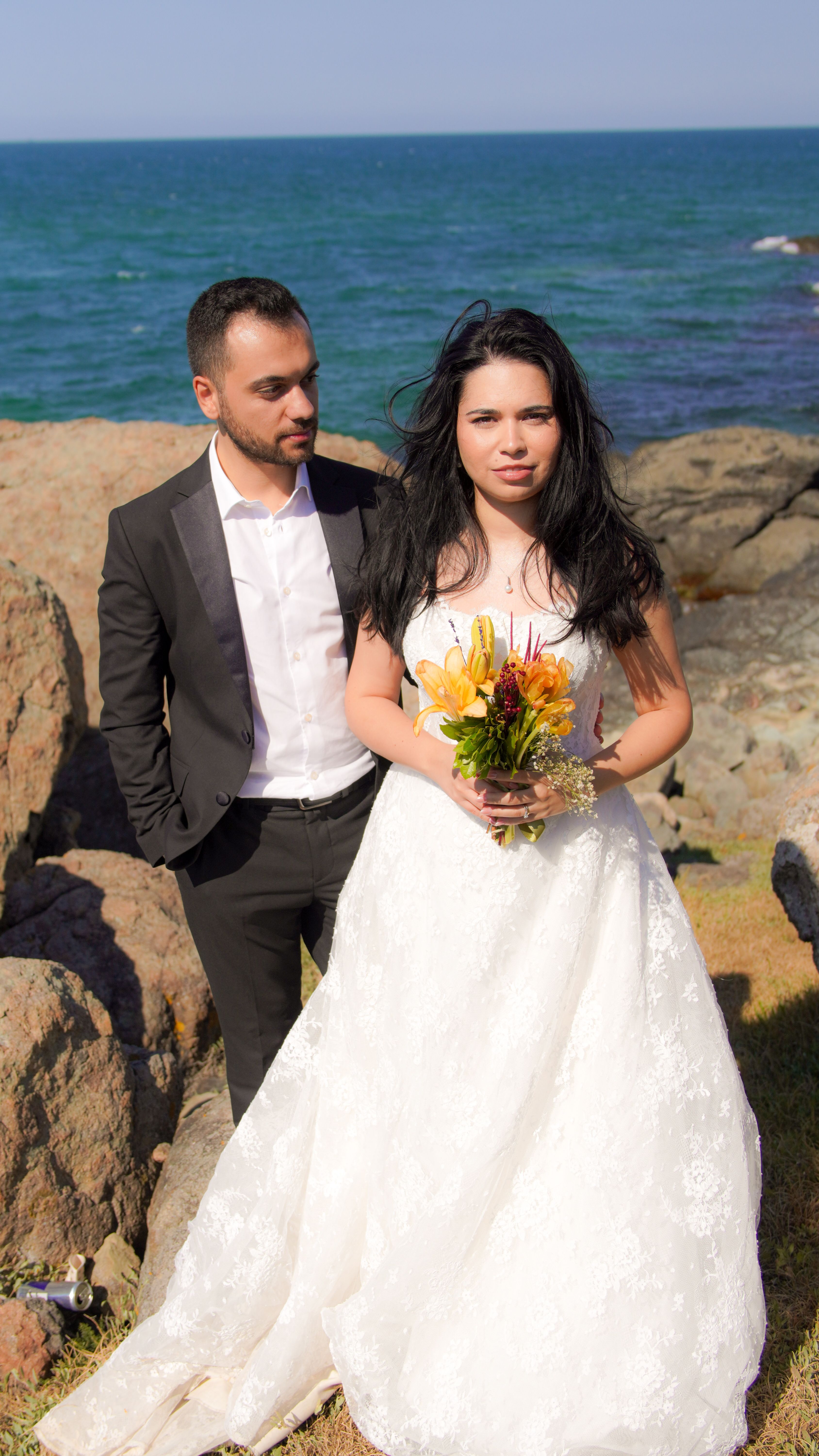 Bride and groom pose by the ocean on a sunny day for a beach wedding.