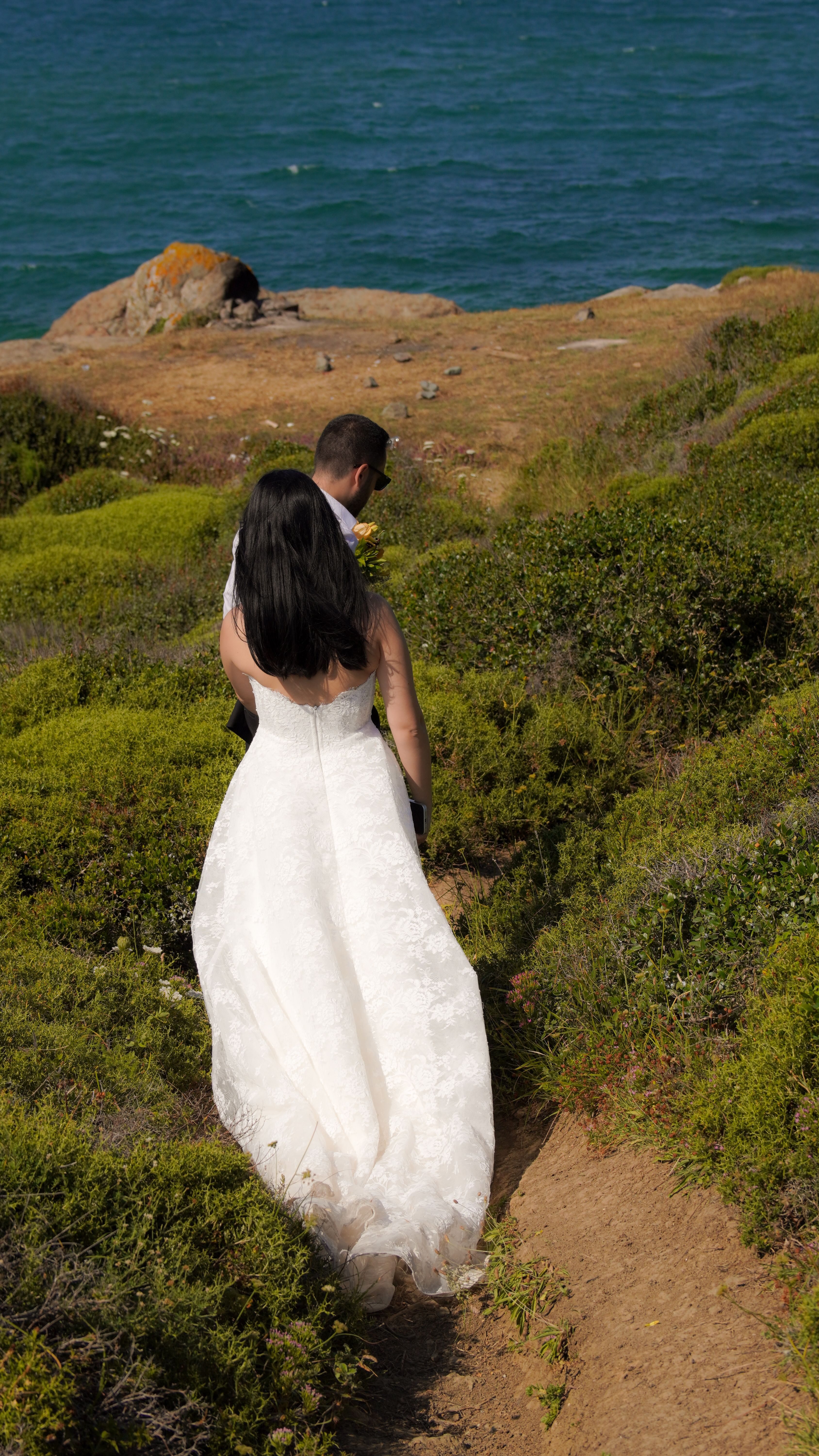 A couple in wedding attire walks along a scenic coastal path, overlooking the ocean.