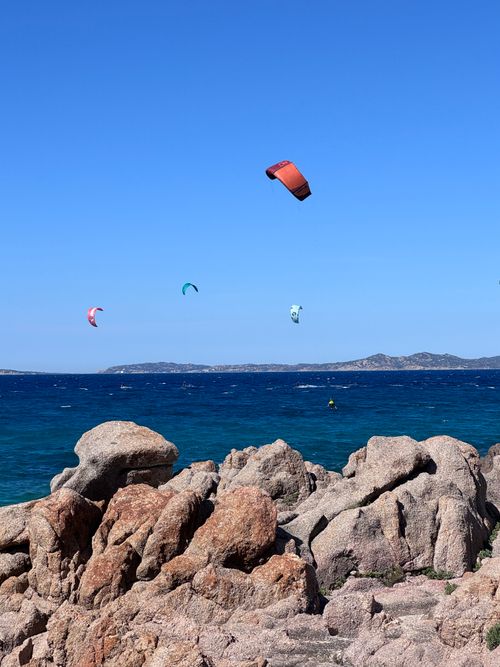 Free Dynamic scene of kite surfers over the blue waters of Sardinia, Italy, against rugged rocks. Stock Photo