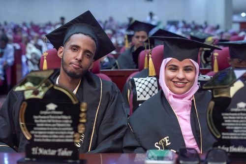 Free Two graduates in caps and gowns celebrate their achievement at a ceremony, showcasing joy and accomplishment. Stock Photo