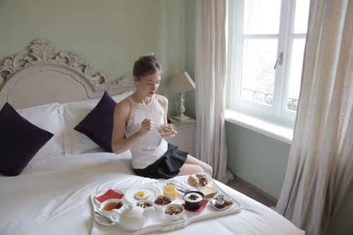 Free Calm female in elegant outfit sitting on bed and eating delicious breakfast served on tray in hotel room with classic interior in morning Stock Photo