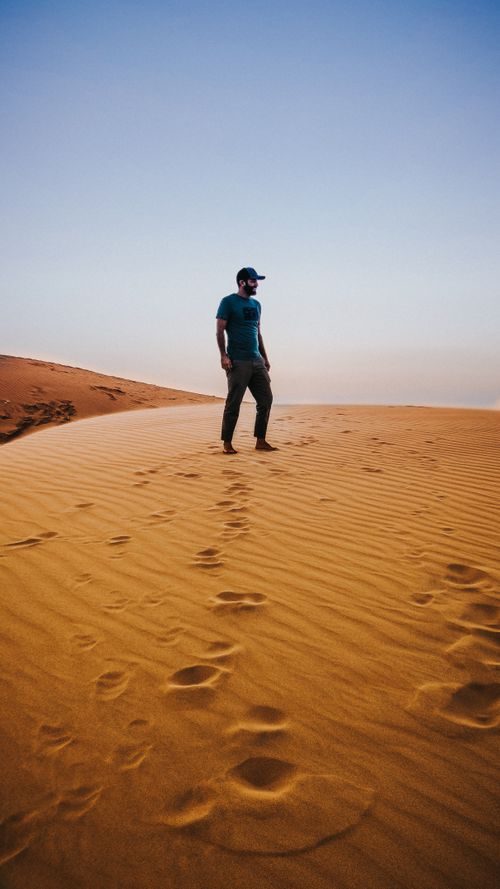 Free Full body of unrecognizable barefooted young male traveler in casual clothes and cap standing on sandy dune of desert against cloudless blue sky Stock Photo