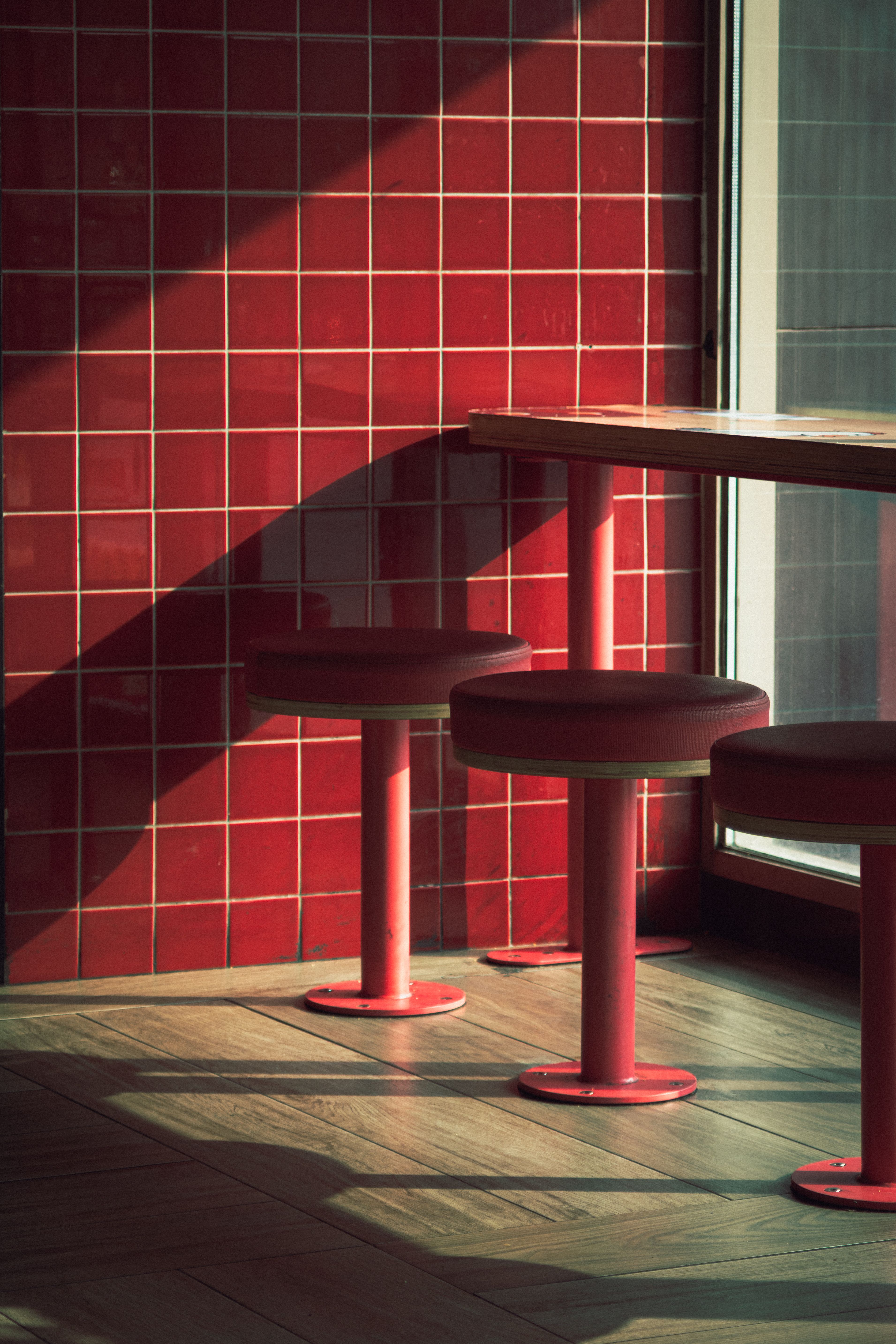 Moody interior shot of red tiled wall and matching stools in sunlight.