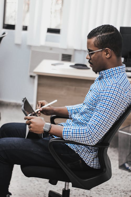 Free Side view of concentrated young African American male employee in casual outfit and eyeglasses using tablet while working in modern office Stock Photo