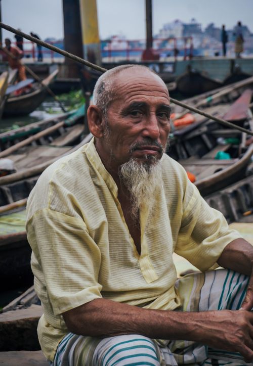Free Elderly man in traditional attire sitting by riverside boats, showcasing cultural and everyday life. Stock Photo