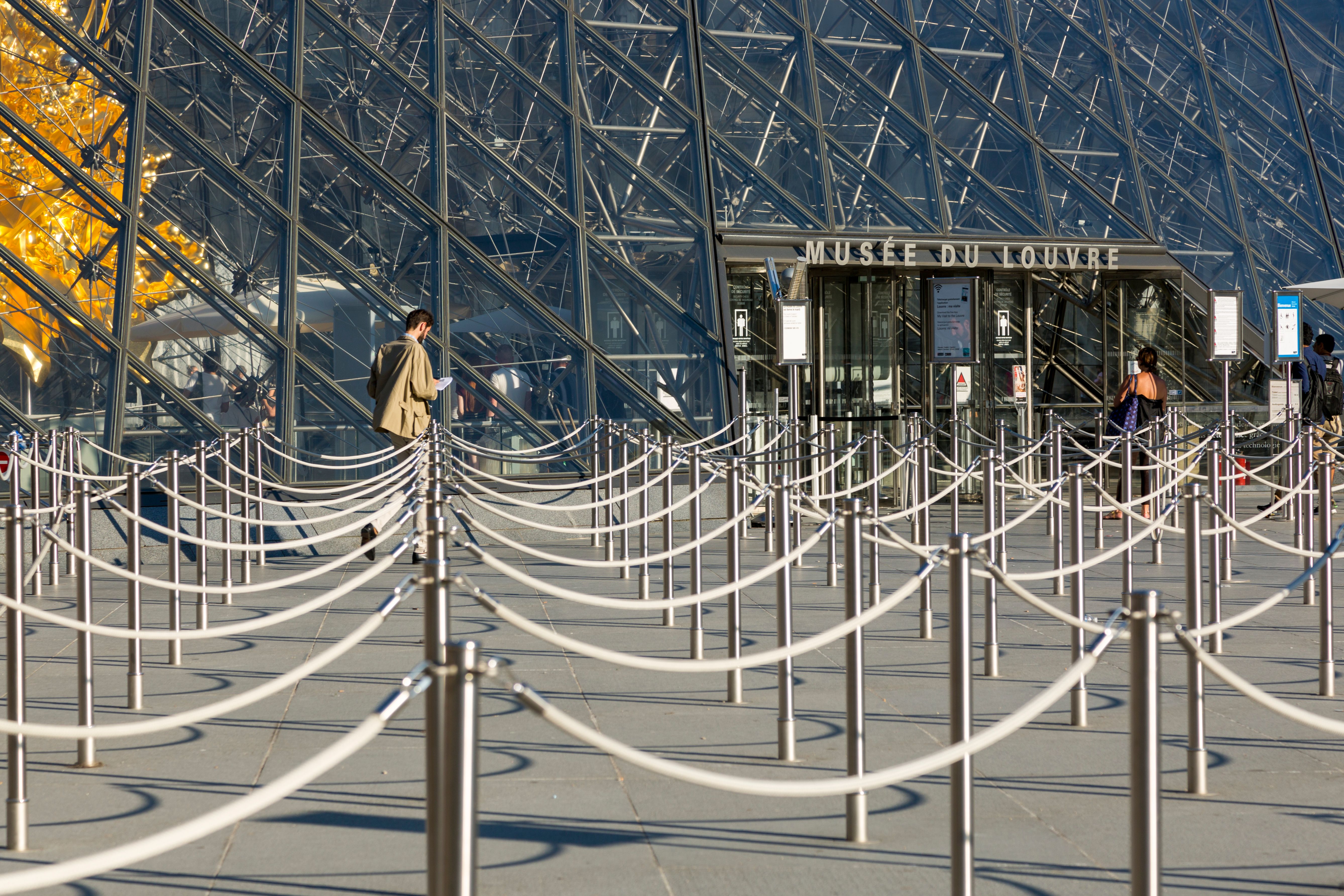 Visitors queued up at the iconic Louvre entrance in Paris, with the glass pyramid reflecting sunlight.
