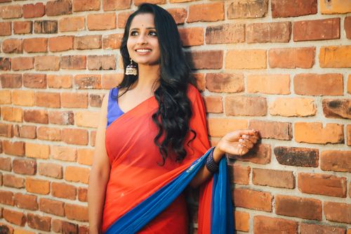 Free Smiling woman in vibrant red and blue sari leaning against a rustic brick wall. Stock Photo