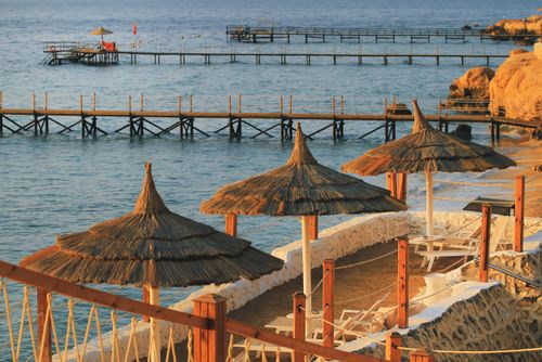 Free Peaceful beachfront scene featuring wooden piers, straw umbrellas, and blue ocean waters at sunset. Stock Photo